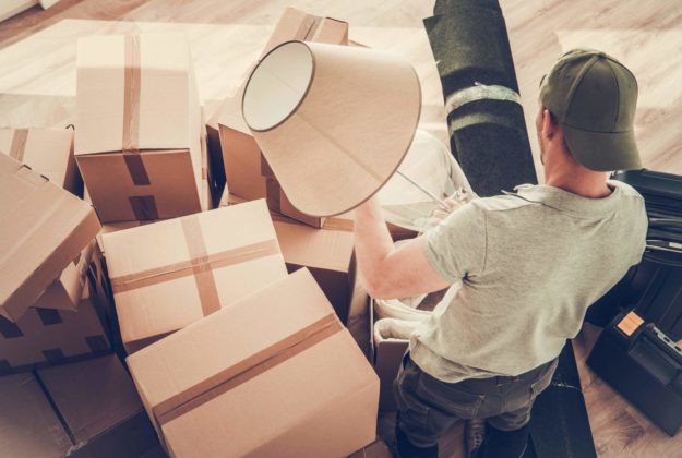 Man Moving Out From His Home. Staying Between Cardboard Boxes Preparing to Pack His Lamp.